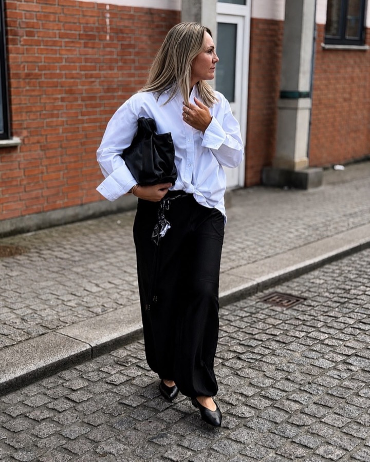Kvinde i hvid oversize-skjorte og lang sort nederdel i en brostensgade